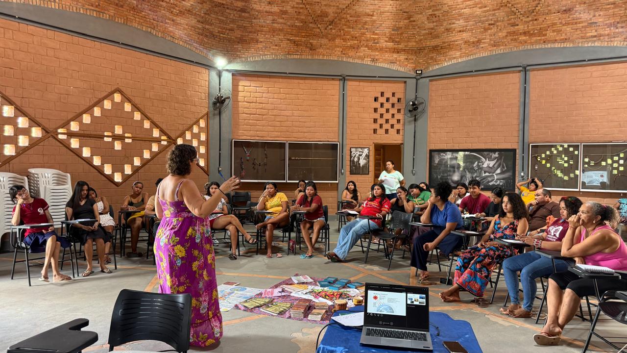 Participantes sentados em círculo acompanham a oficina formativa conduzida pela professora Fabrina Furtado em amplo espaço de alvenaria na Aldeia Piçarra Preta. Ao centro, livros e materiais educativos estão dispostos no chão junto a um computador com projeção de apoio visual.
